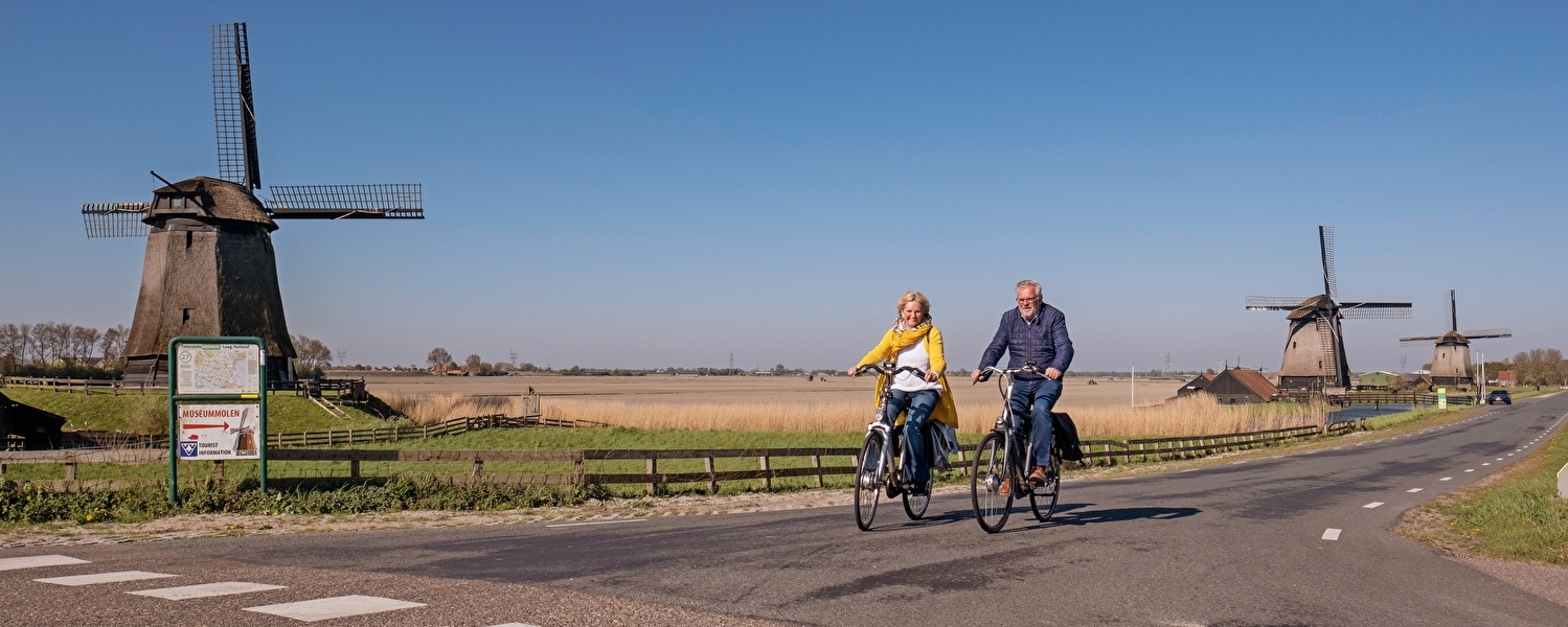 Stel fietst door het Hollandse landschap bij Alkmaar, langs traditionele windmolens en uitgestrekte velden onder een strakblauwe lucht.