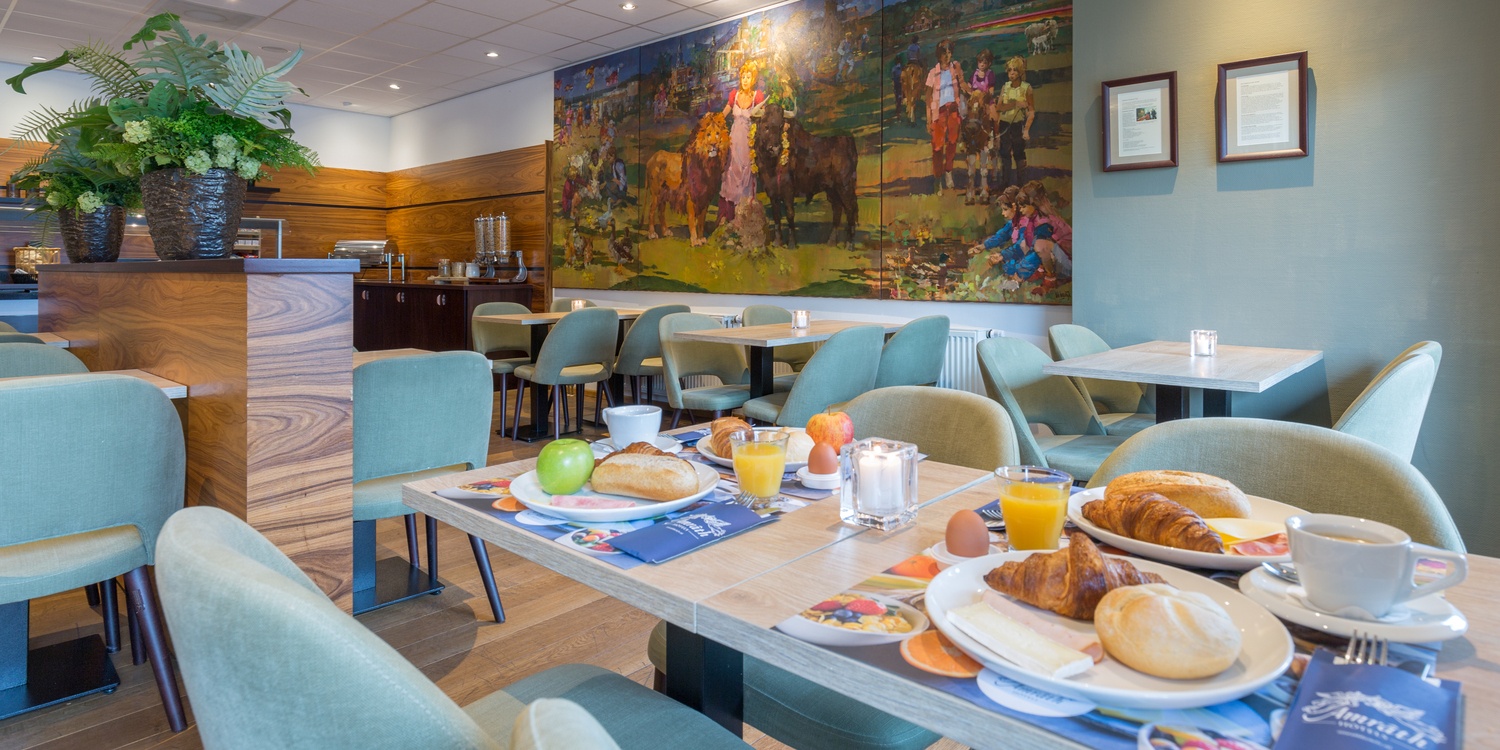 Restaurant gedeelte van Amrâth Hotel Alkmaar met houten vloeren, groene gestoffeerde stoelen en een kleurrijk schilderij aan de muur, wat een warme ambiance creëert.